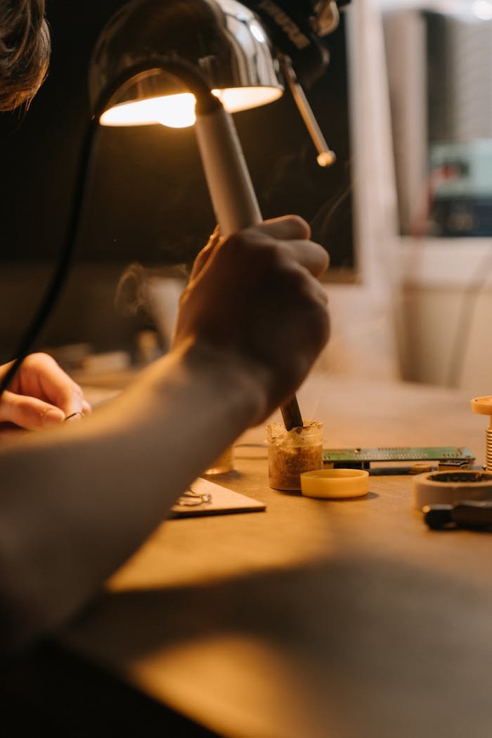 A detailed view of a hand soldering electronic components under a lamp on a workbench.